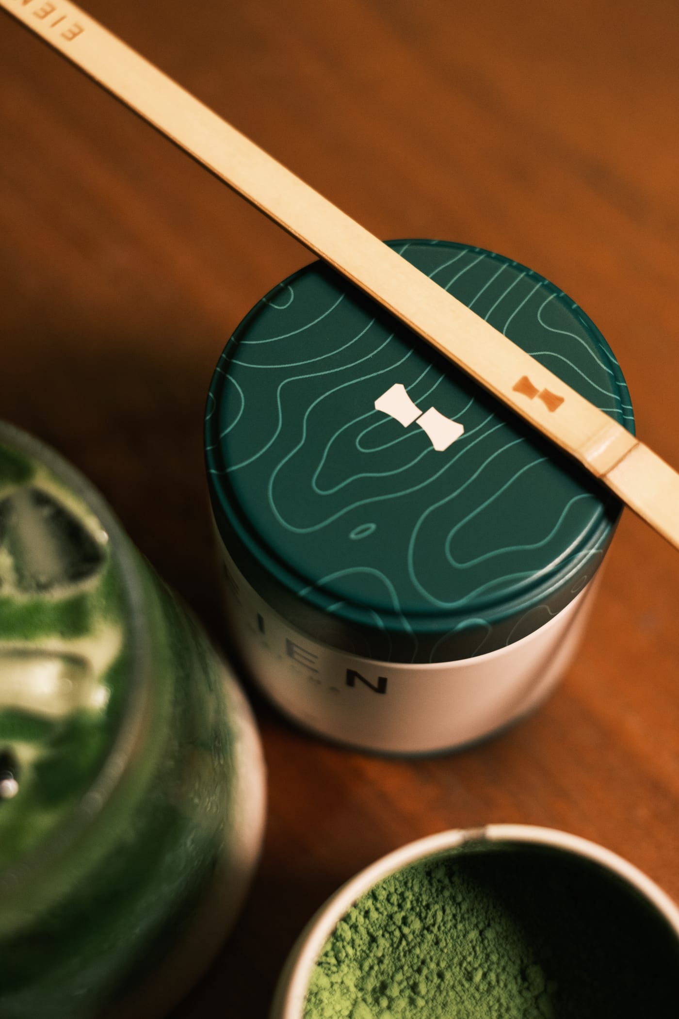 A tin of EIEN matcha on a dark wood table, a chashaku resting across its lid beside a bowl of sifted powder